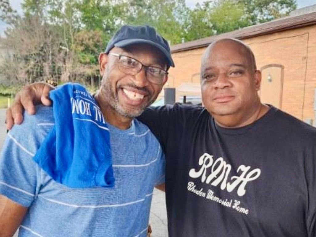 Two men smiling, arm around each other. One in a blue hat and shirt, the other in a black shirt. Outdoors.