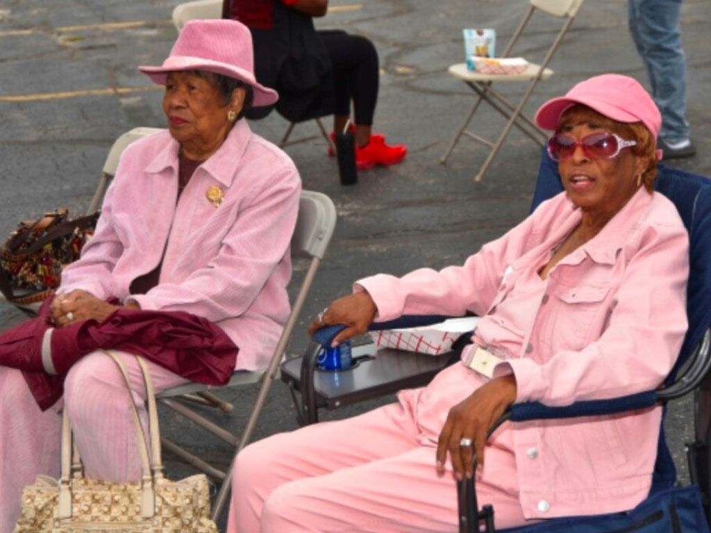 Two older women in pink outfits sit outside, one in a hat, the other in sunglasses.