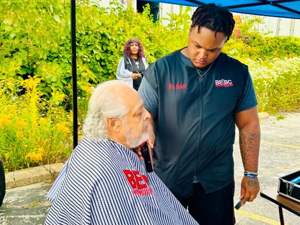 Man getting a haircut outside under a blue canopy. Barber wearing black vest cuts client's hair.