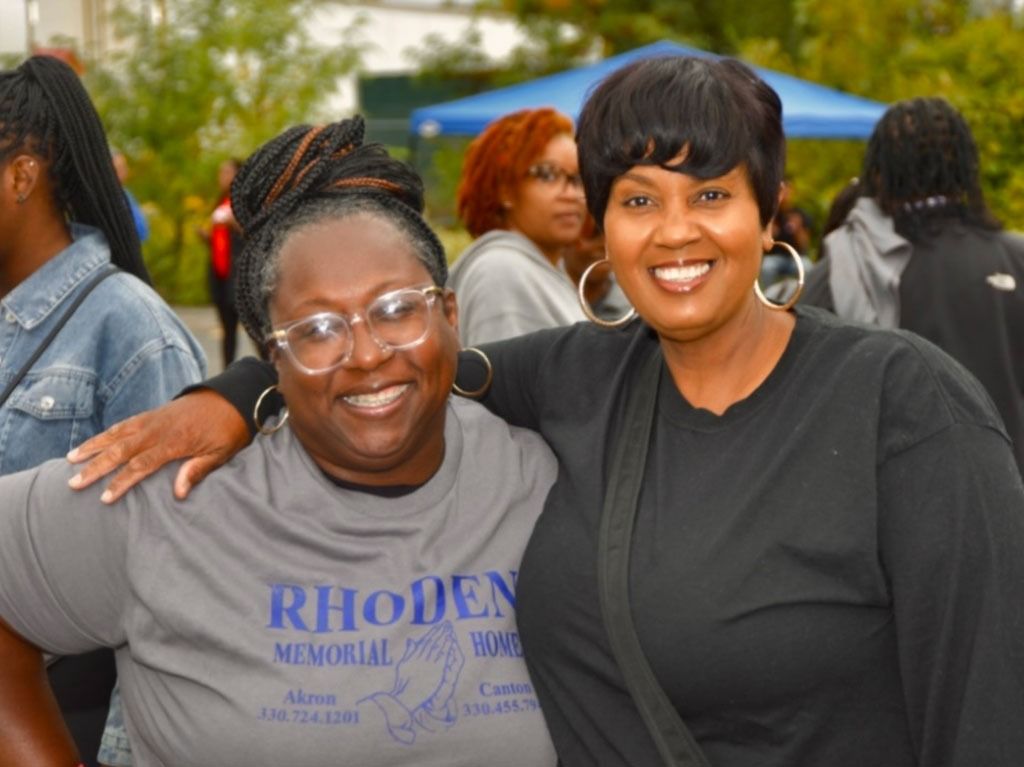 Two smiling Black women pose for a photo at an outdoor event; one has her arm around the other.