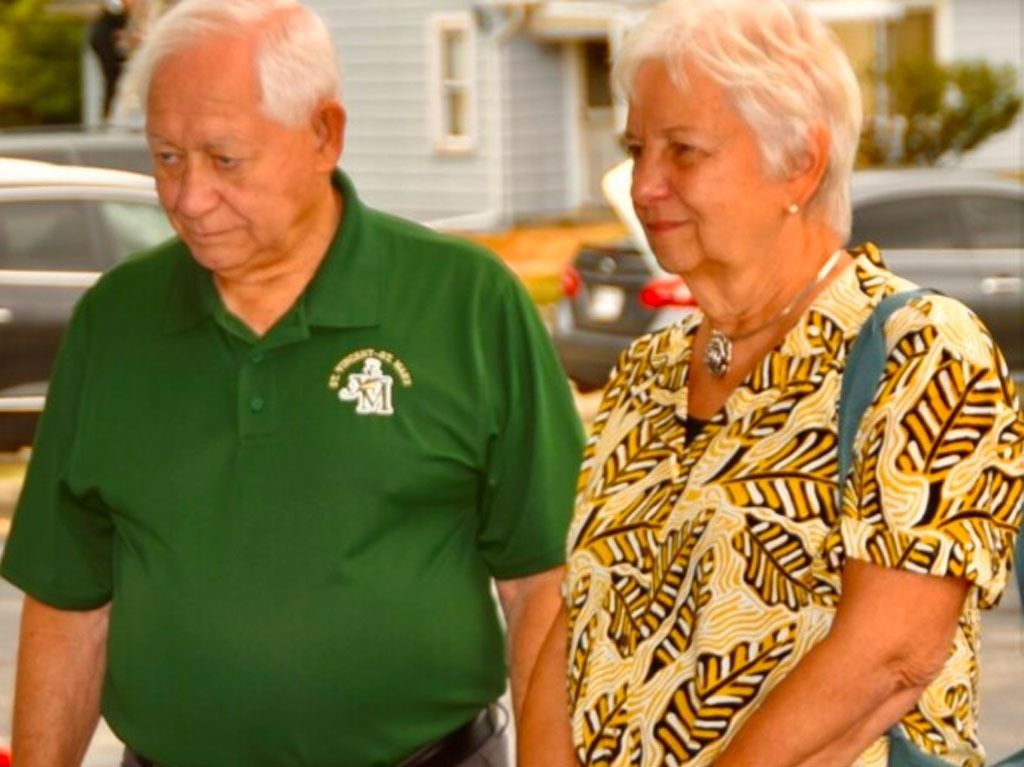 Elderly couple in green and patterned shirt outdoors. Man looks down, woman smiles, background cars.