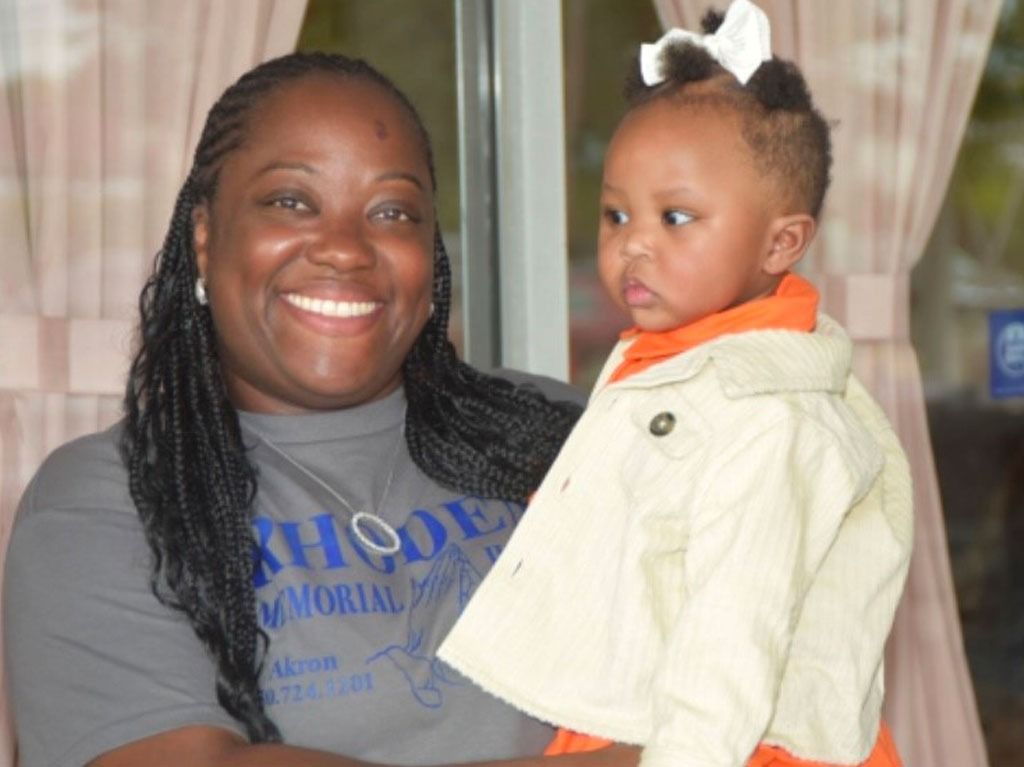 Woman with braids smiles, holding a baby with a white bow. They're indoors, near a window.