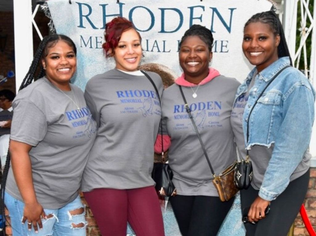 Four women pose in front of a memorial banner. Three wear gray shirts, one in a denim jacket.