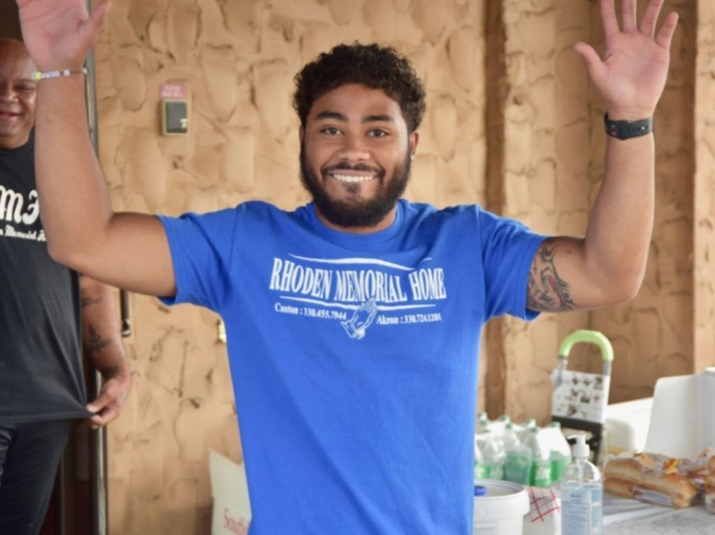 Man with arms raised, smiling, wearing a blue t-shirt that says