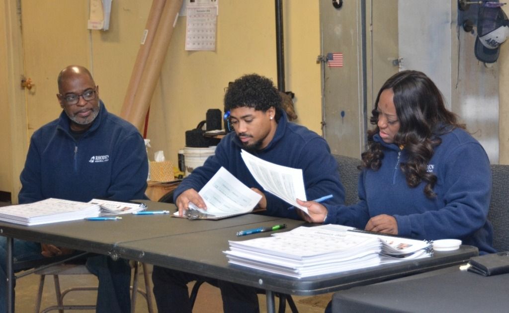 Three people in blue sweatshirts review papers at a table in an industrial setting.