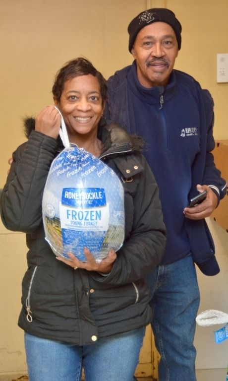 Woman holding frozen turkey, smiling, with man behind her, both indoors.