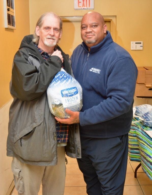 Two men holding a frozen turkey; one smiles, the other wears a blue work uniform. Indoor setting.