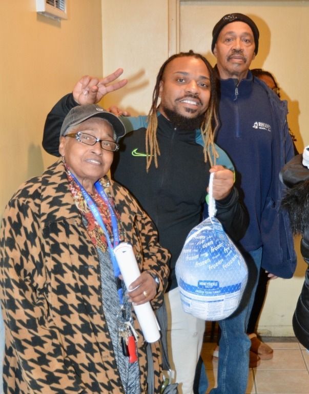 Three people pose, smiling, holding a wrapped turkey. Woman in houndstooth coat, man in black jacket, another in blue.