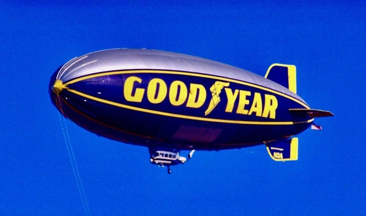 Goodyear blimp with the Goodyear logo, flying in a clear blue sky.