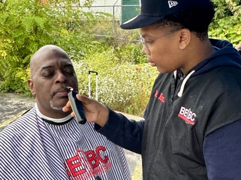 A barber shaves a man's facial hair outdoors. Both are Black; the barber wears a hat and jacket.