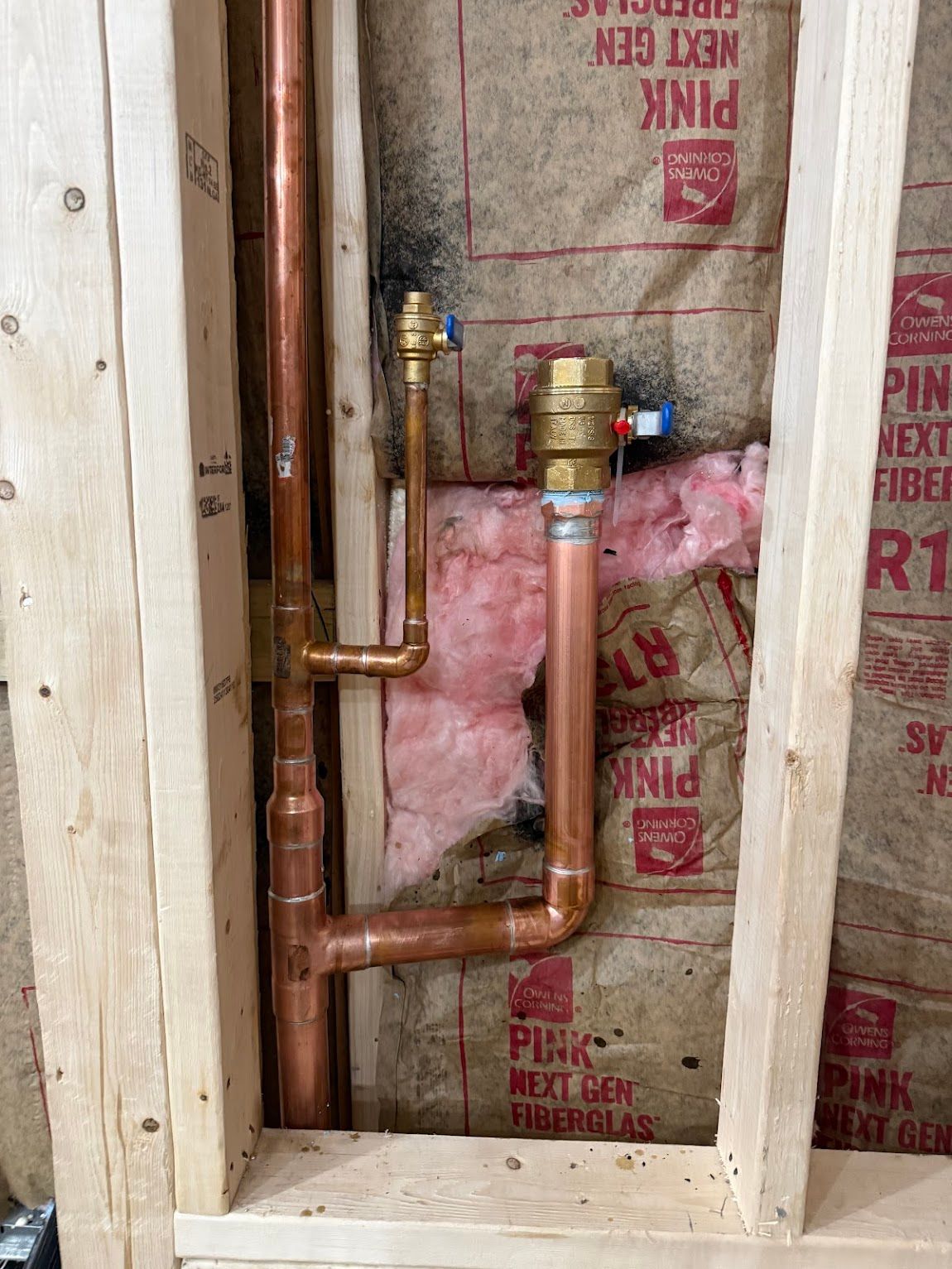 Copper plumbing pipes and brass valve inside wall framing with pink insulation.