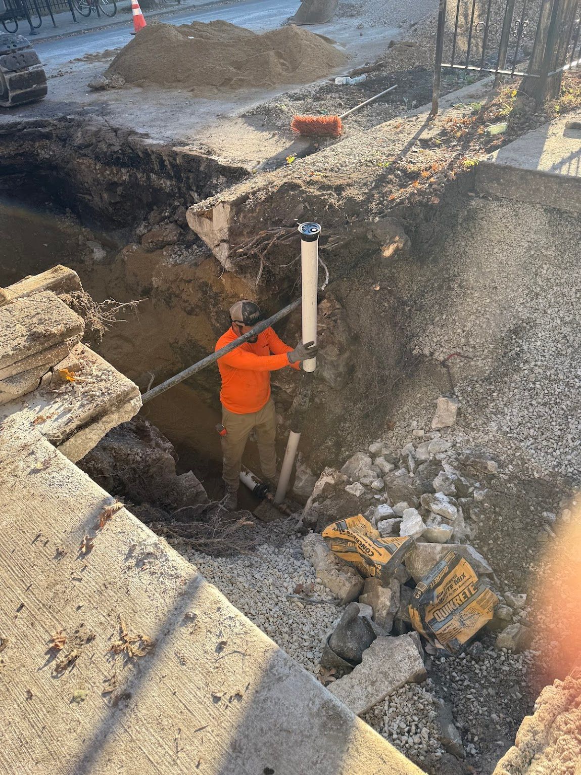 Worker in orange jacket standing in a deep trench at a construction site, holding a pole near a white pipe.
