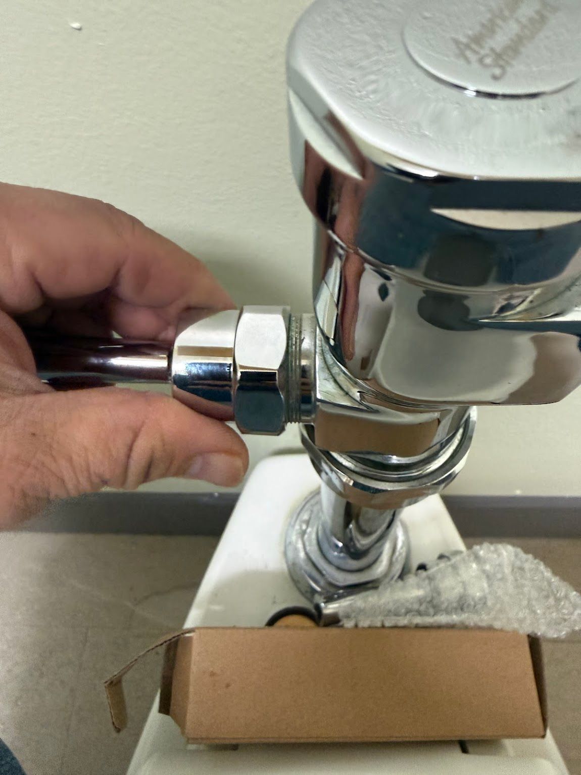 Hand turning a chrome valve on a toilet flush mechanism with a cardboard box below it.