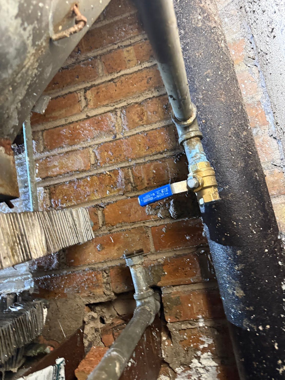Close-up of a brick wall with exposed plumbing pipes and a blue-handled shutoff valve.