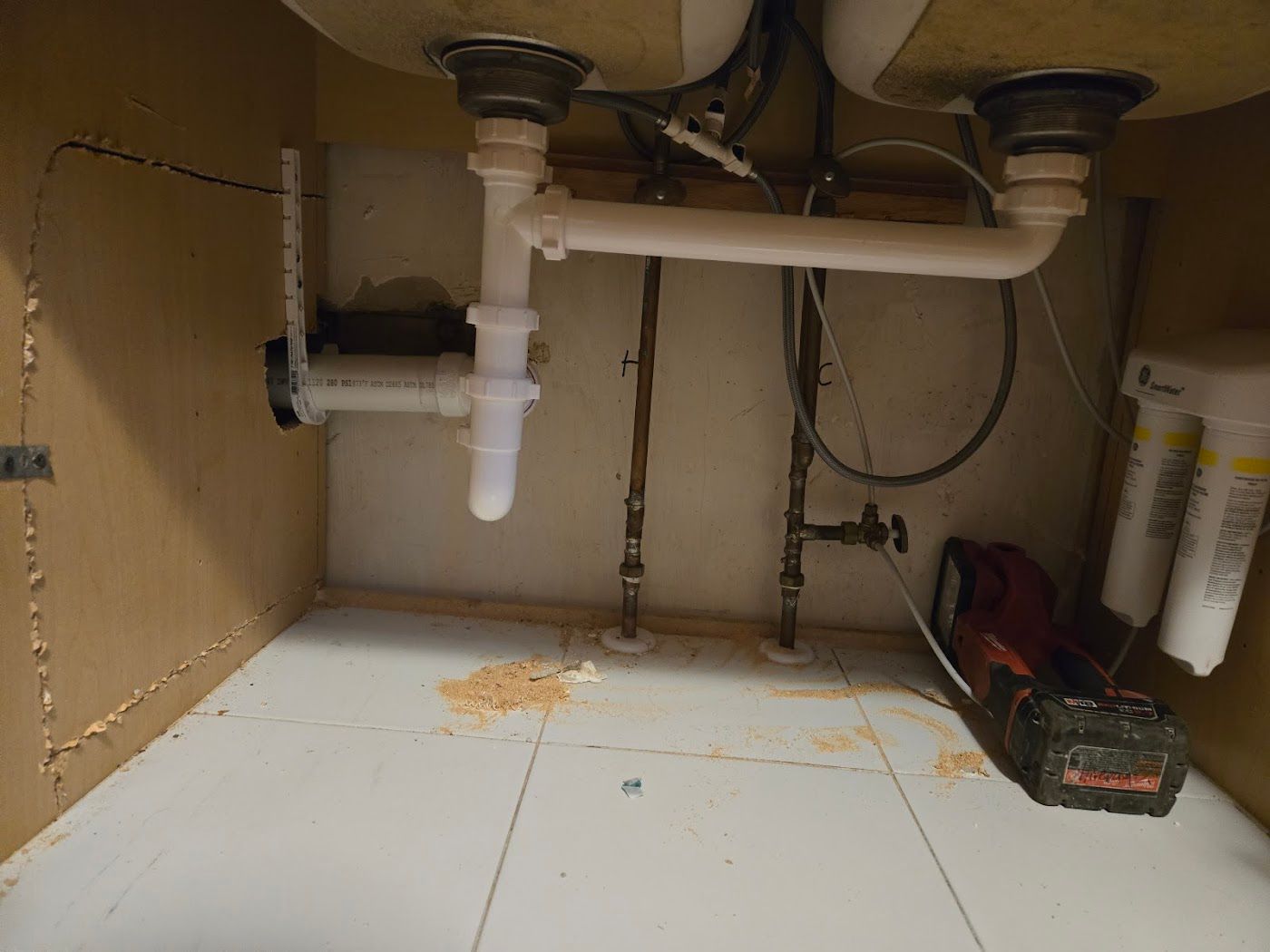 Under-sink plumbing and storage area with white pipes, scattered debris, and a red toolbox on the right