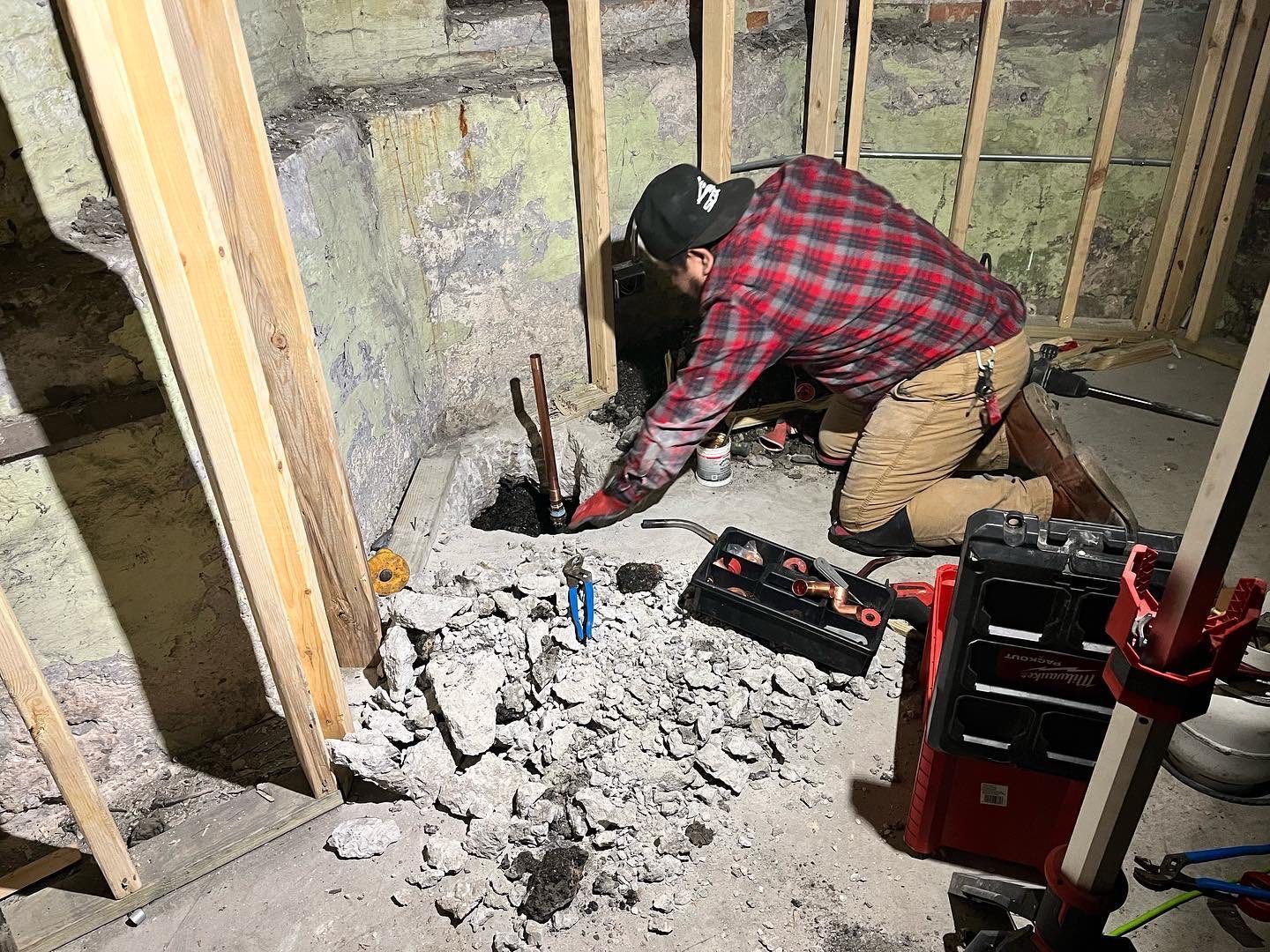 Worker kneeling amid insulation debris, repairing a basement wall with tools and exposed studs nearby