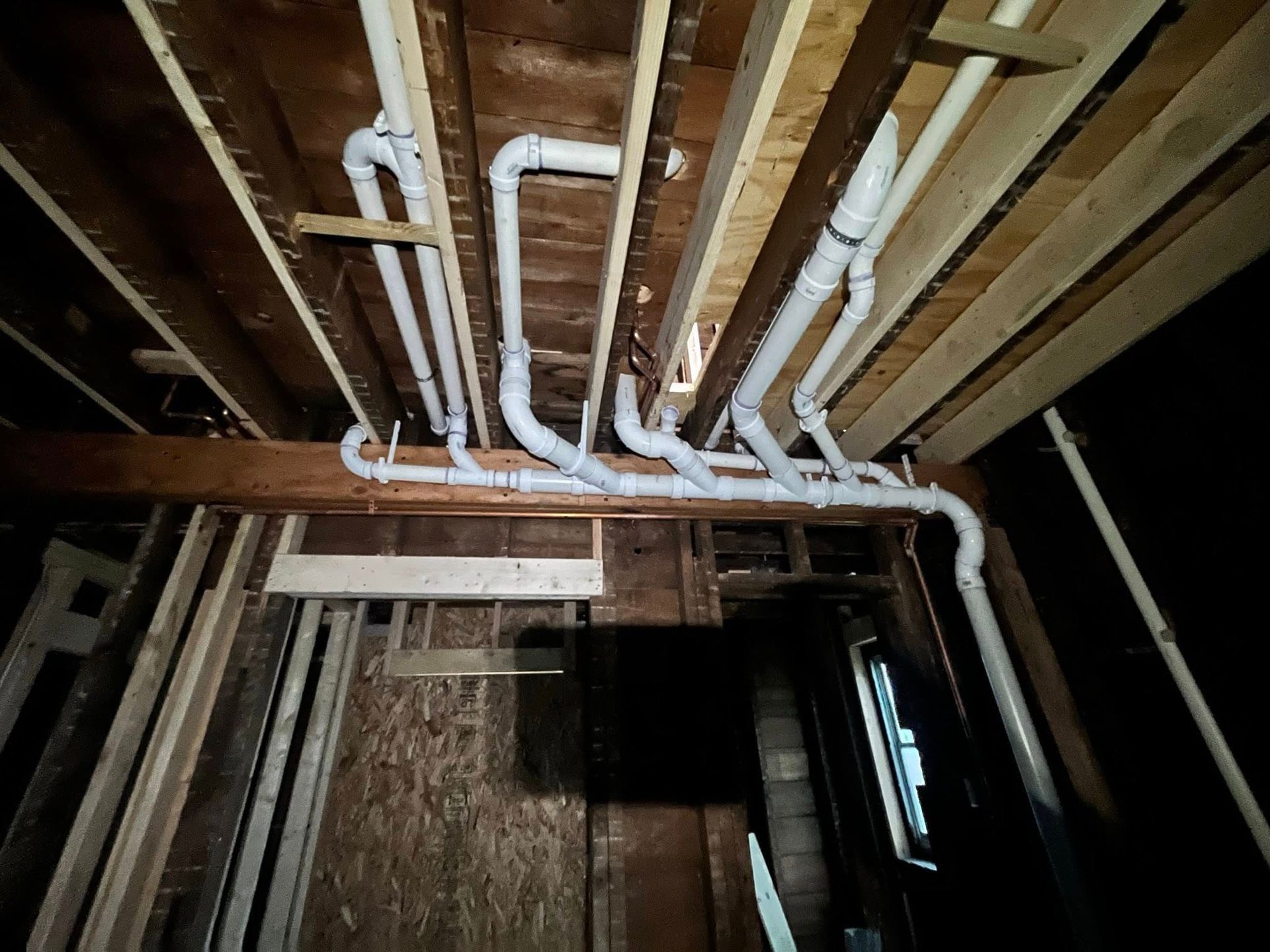 Exposed white PVC pipes and wooden ceiling framing in an unfinished basement or crawlspace.