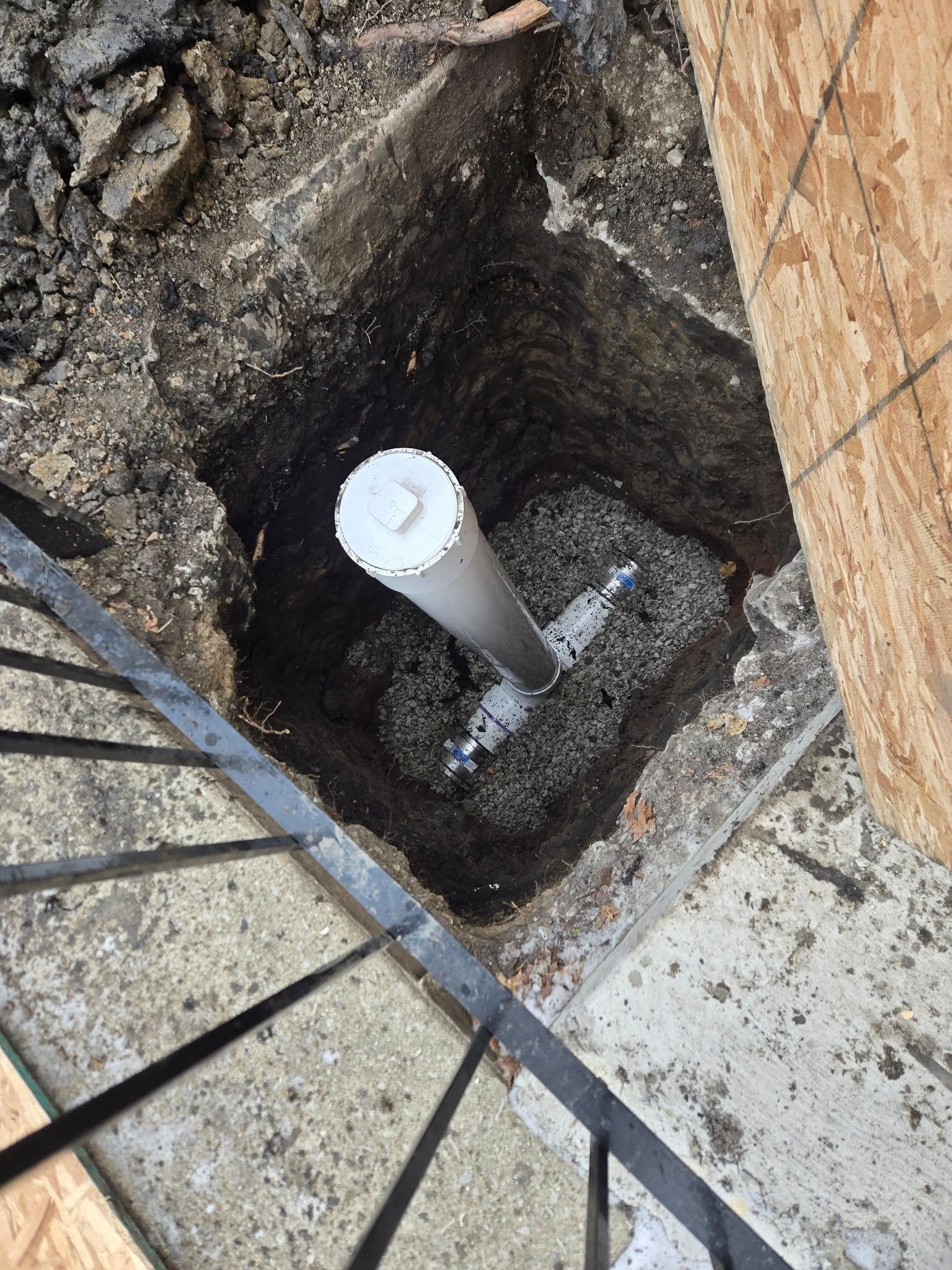 Open utility pit with a white PVC pipe and dirt around it next to a wooden wall and metal railing.