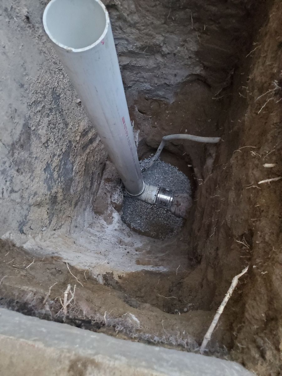 Excavated hole with a white PVC pipe and a small metal fitting at the bottom
