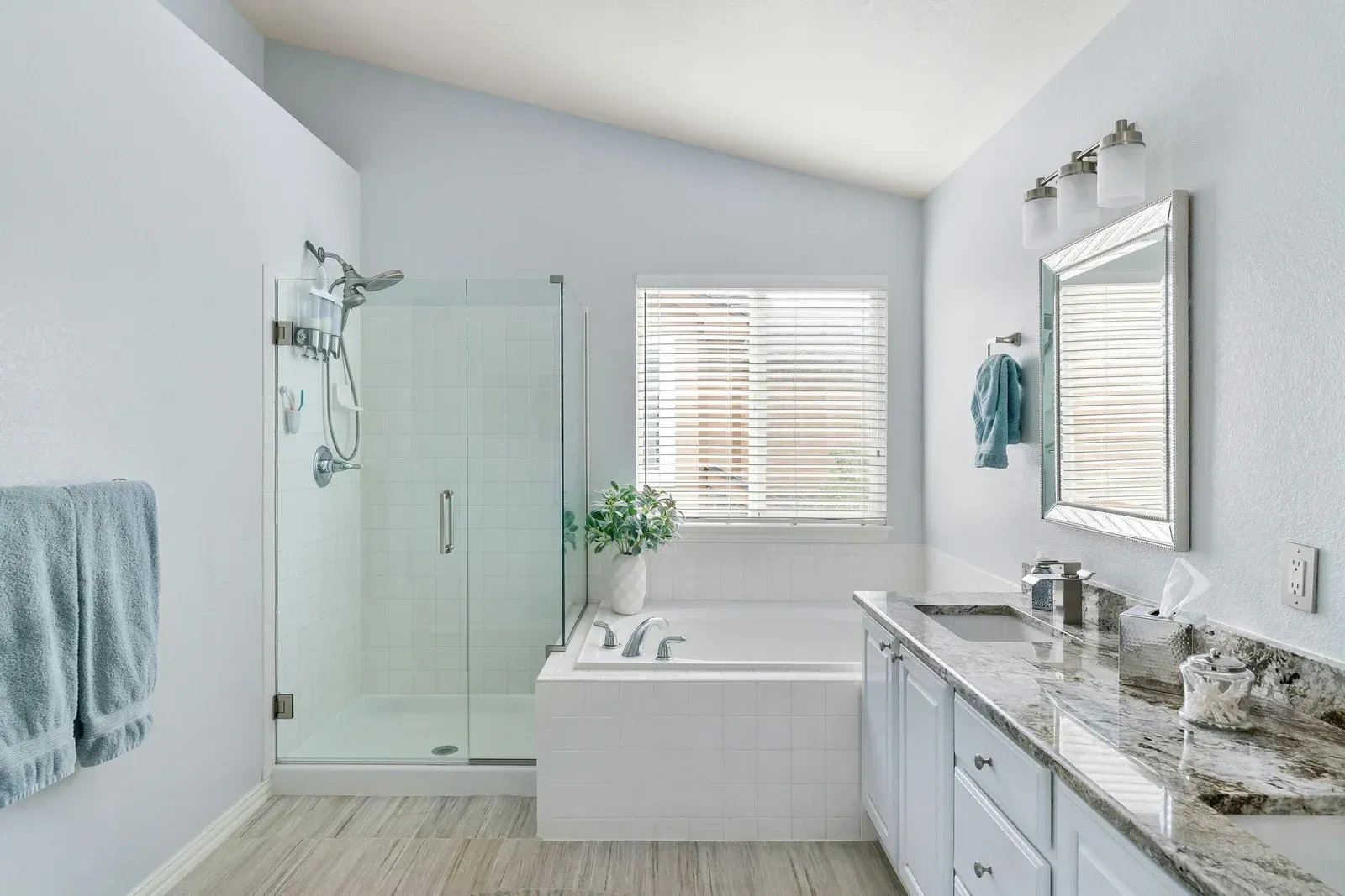 Bright modern bathroom with glass shower, bathtub, double vanity, and window blinds