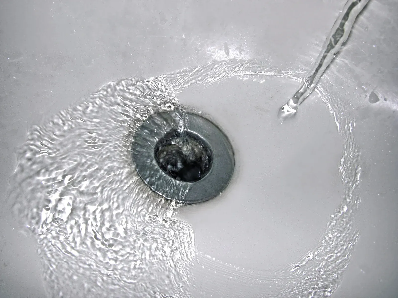 Close-up of water flowing into a white sink drain with a metal stopper.
