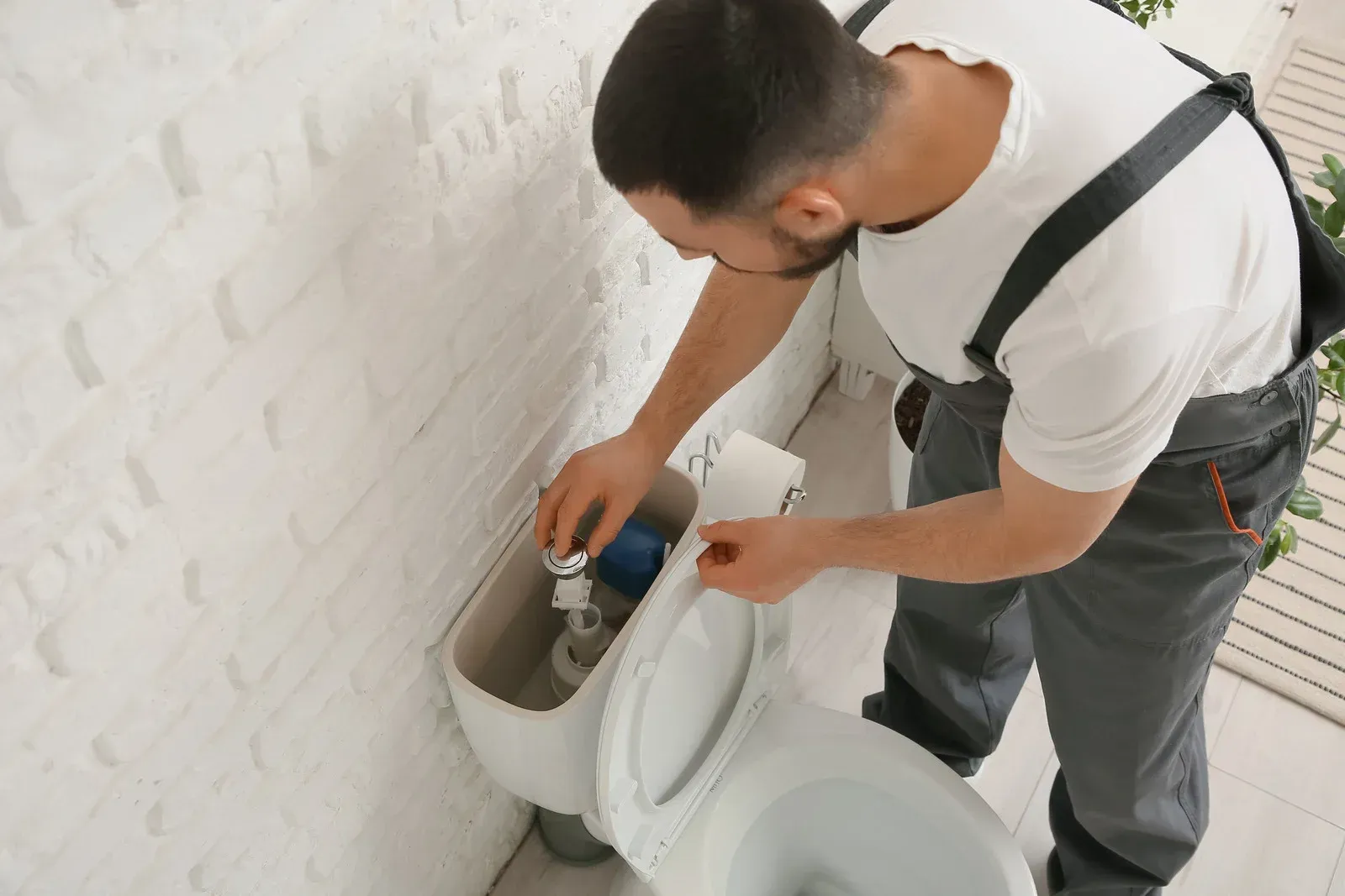 Man repairing a toilet tank in a white bathroom, using a tool beside the open toilet seat