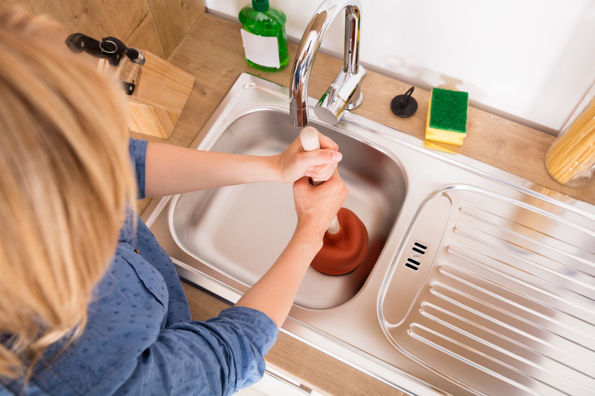 High Angle View Of Woman Using Plunger In Blocked Kitchen Sink To Unclog Drain