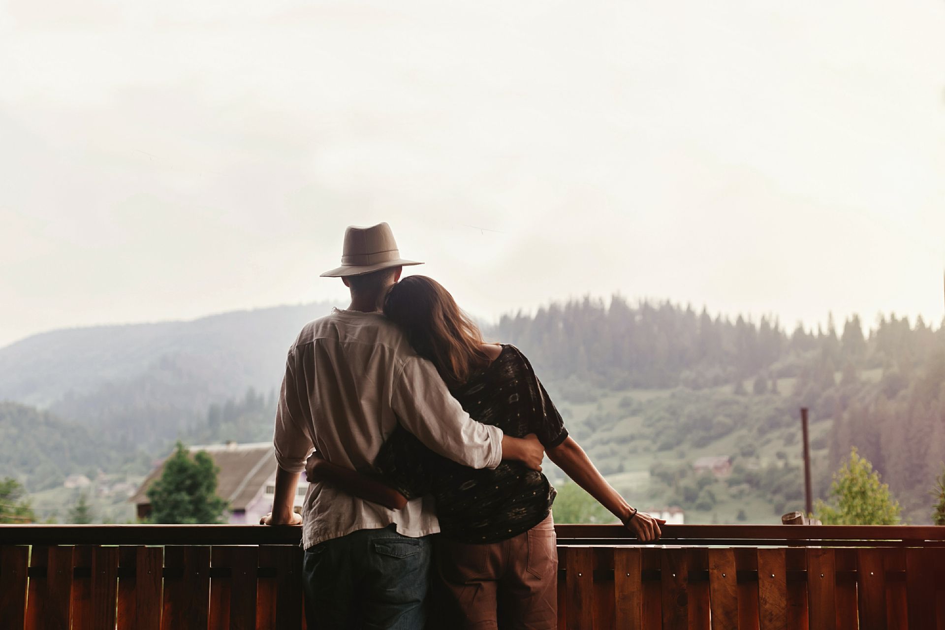 Couple on a wooden balcony overlooking a mountain range. Man in a hat has his arm around woman.
