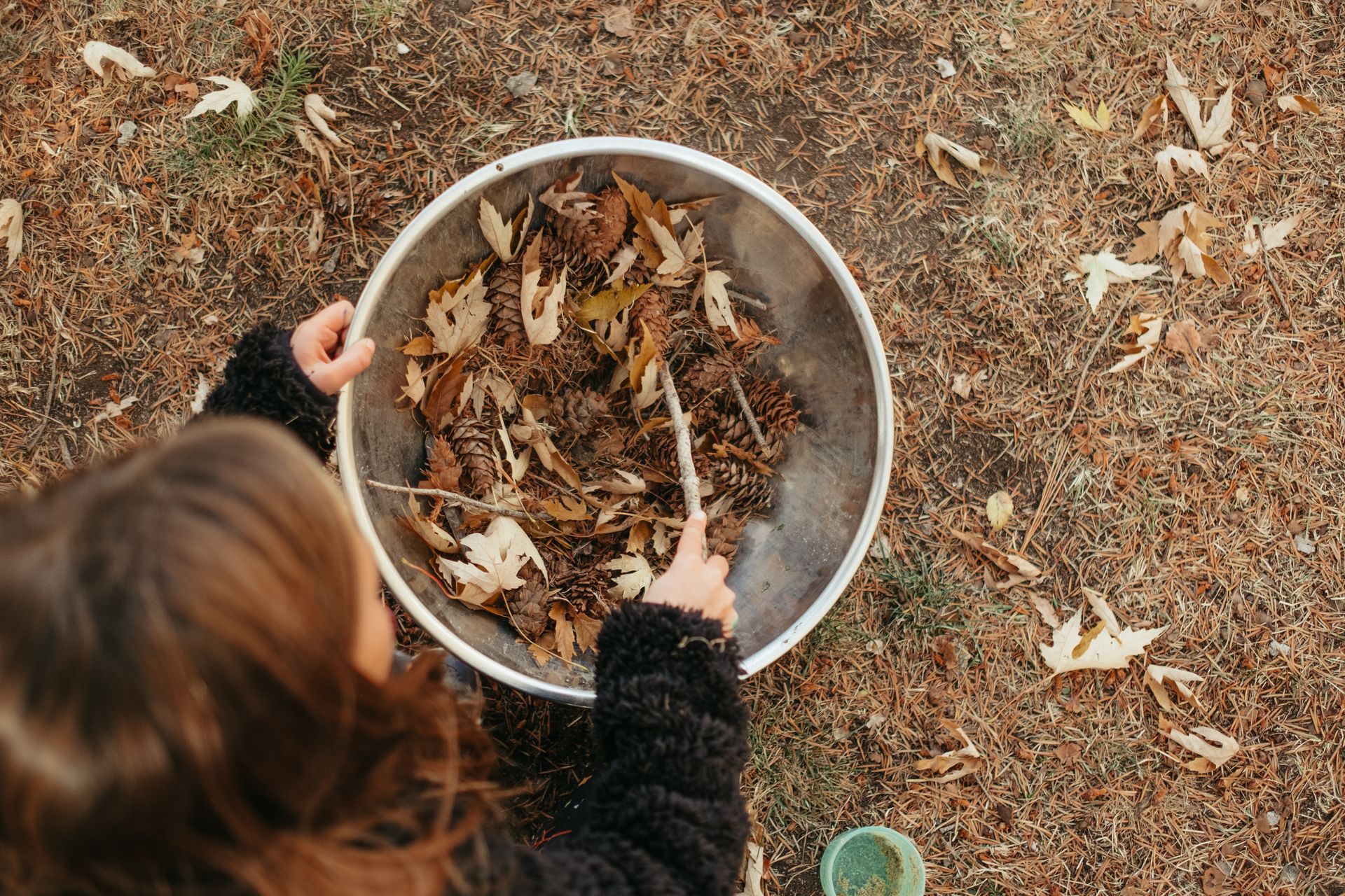 A person holding a stick while mixing dry autumn leaves in a large, round metal bowl on the ground.