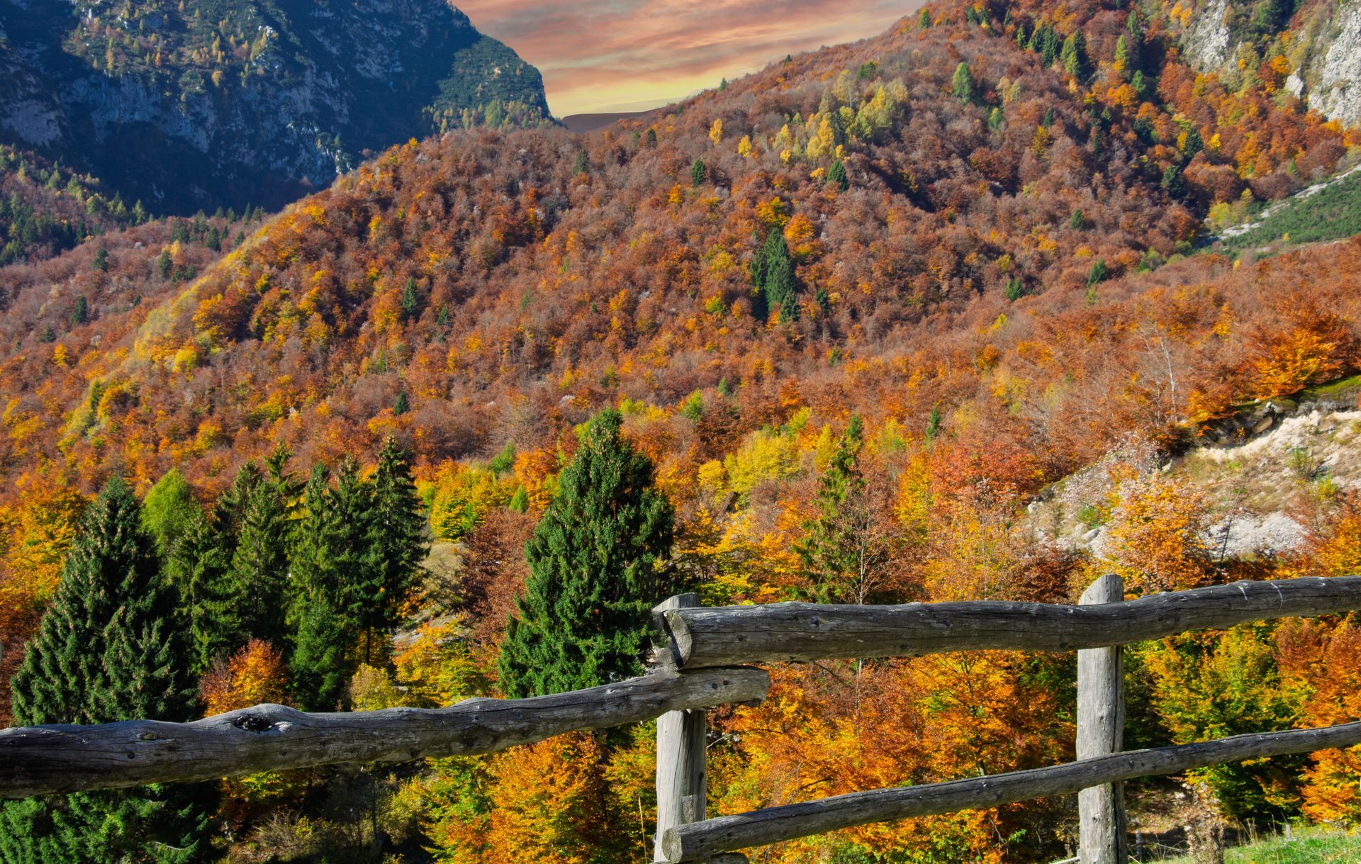 Autumnal mountain landscape with vibrant orange, yellow, and red foliage; a wooden fence in the foreground.