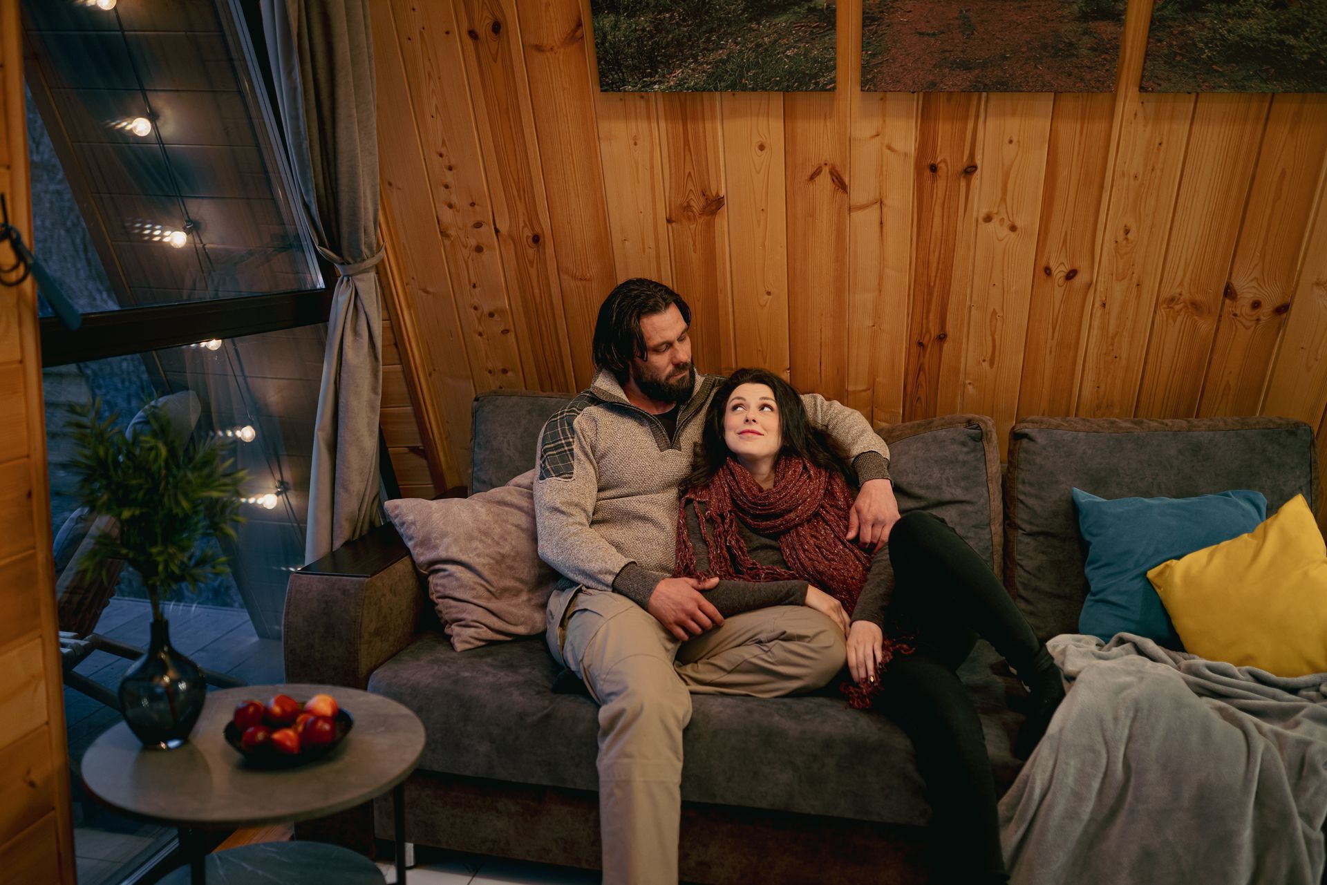 Couple cuddling on a gray sofa inside a wooden cabin. Fairy lights, side table with apples.