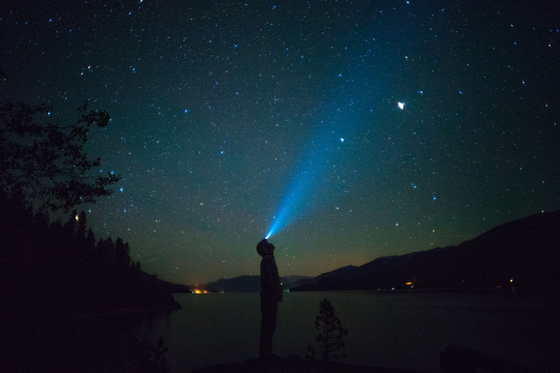 Person shines a flashlight into the night sky filled with stars, standing by a lake with hills in the background.