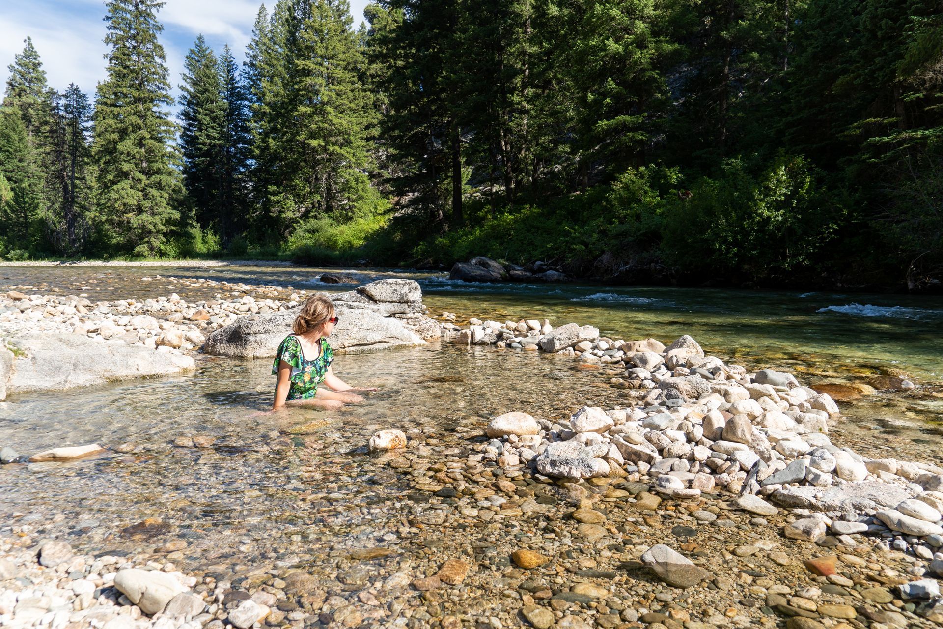 Person seated in a shallow, rocky hot spring by a river, surrounded by trees.
