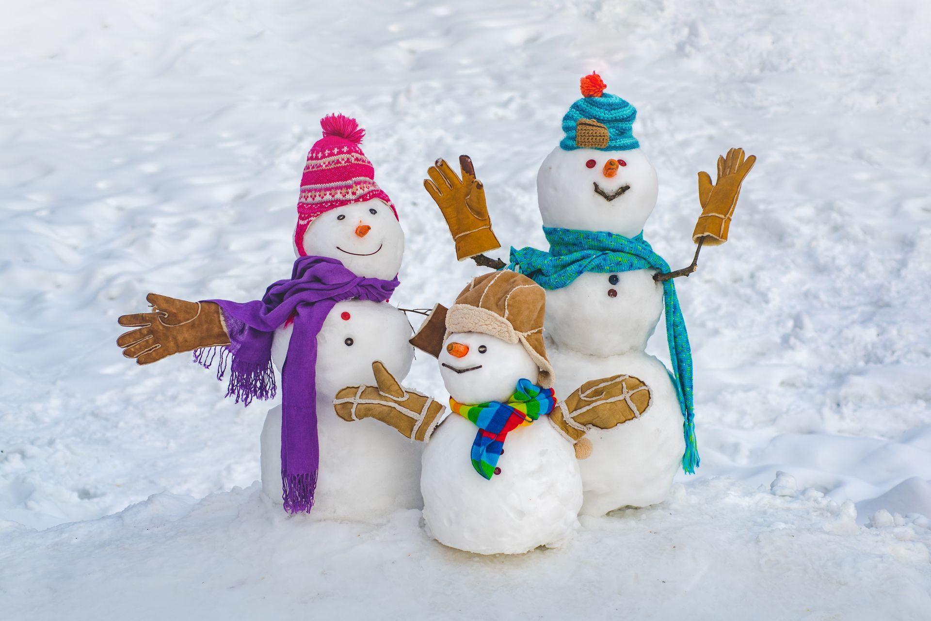 Three snowmen wearing colorful hats, scarves, and gloves stand together in a snowy field.
