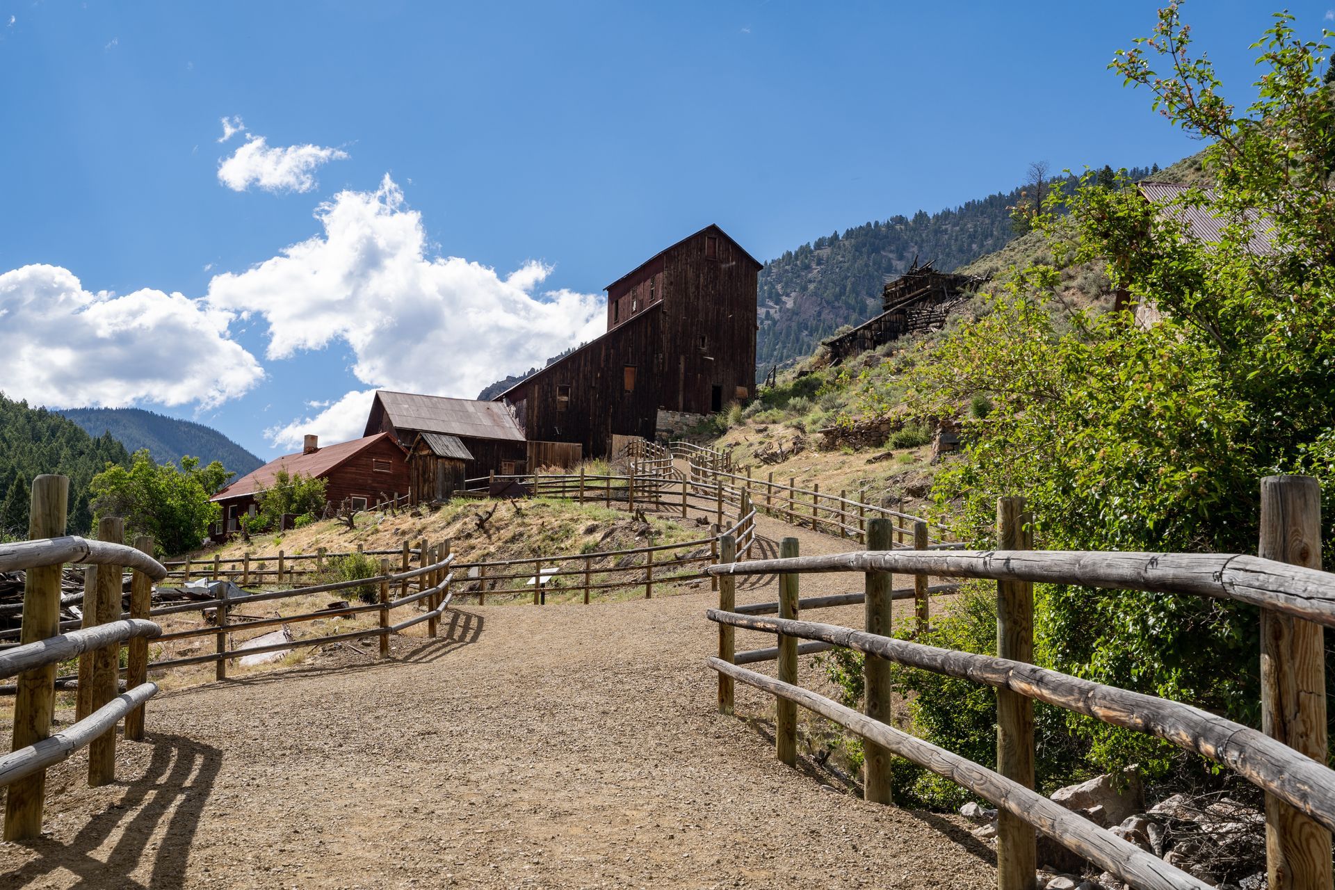 Rustic wooden buildings and fence on a gravel path, under a blue sky, leading to the hills.