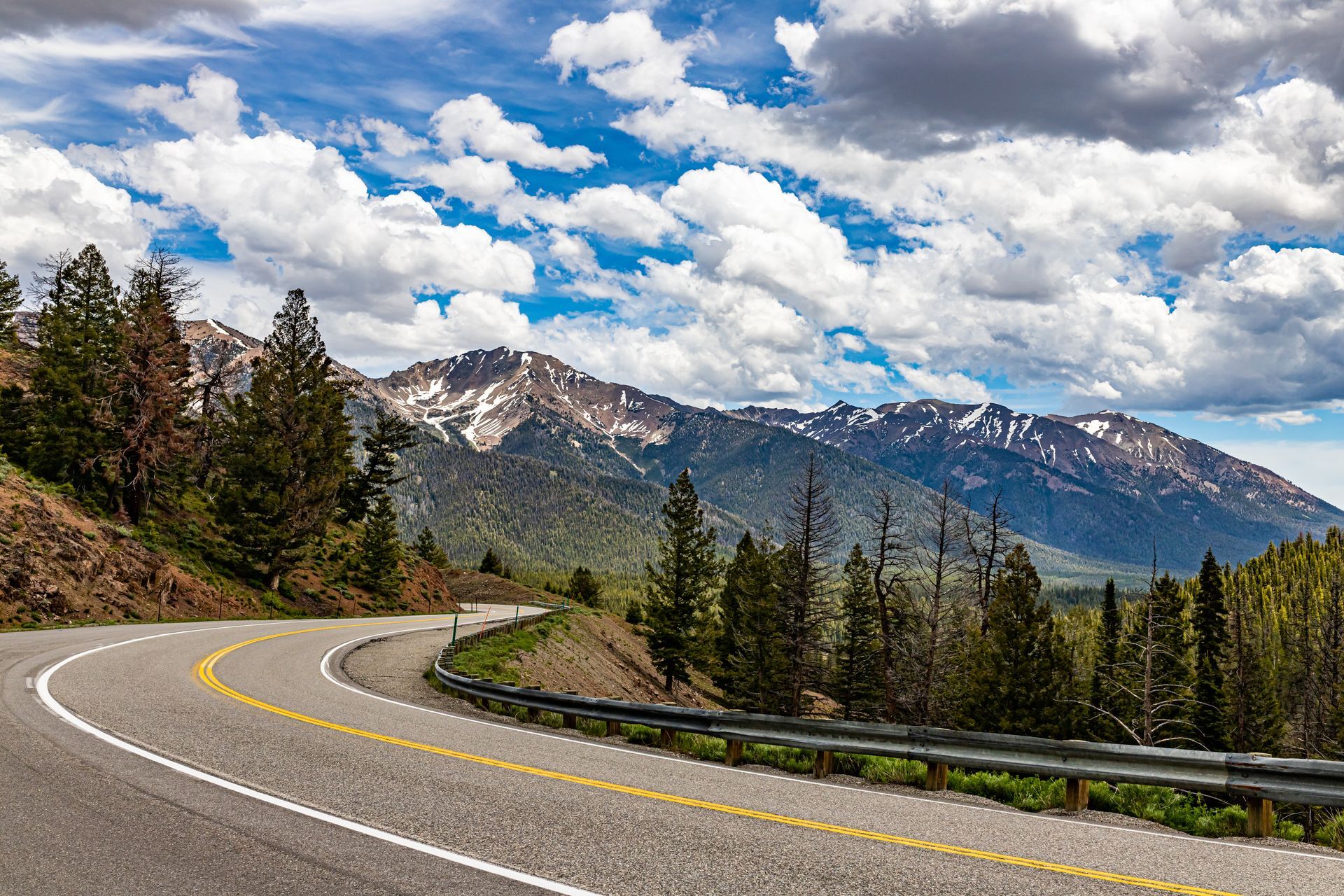 Curving mountain road with evergreen trees, snow-capped peaks, and a cloudy blue sky.