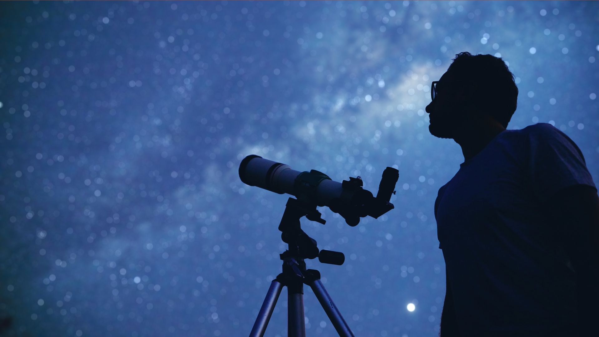 Silhouette of person gazing at a starry night sky with a telescope on a tripod.