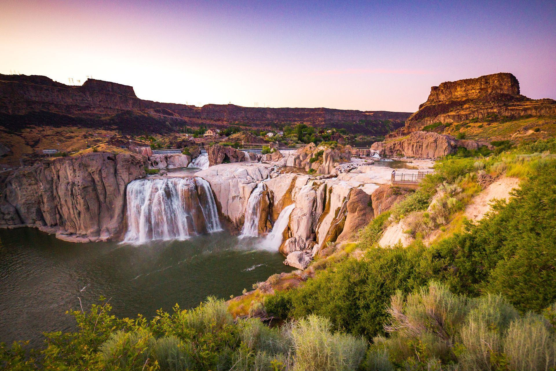 Waterfall cascading over a rocky cliff into a river, surrounded by green shrubs and desert mesas at sunset