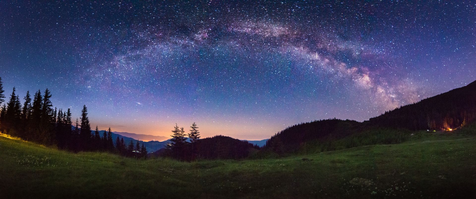 Night sky over a grassy field and dark mountains with the Milky Way visible.