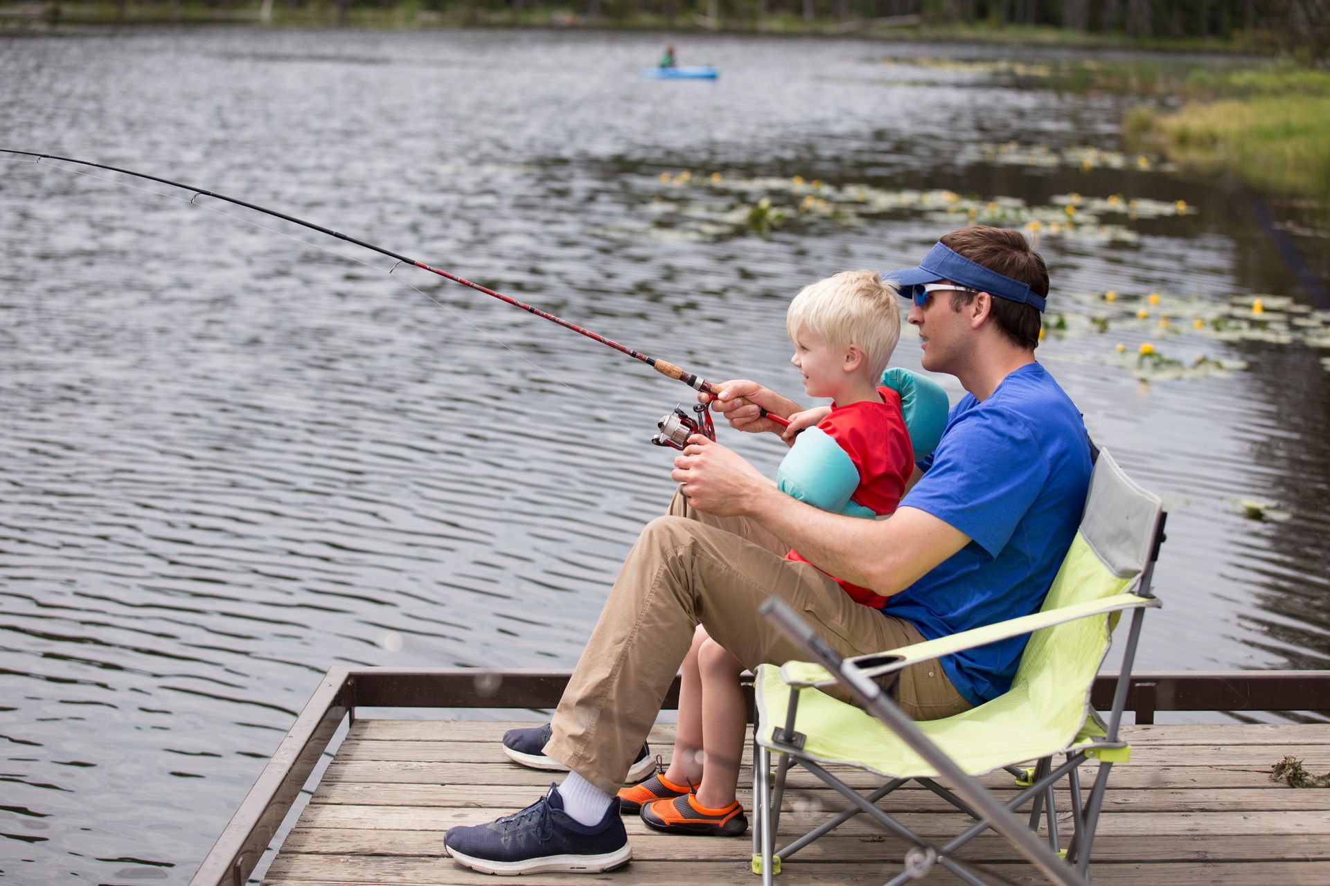 A person wearing a blue shirt and visor sits on a folding chair on a wooden dock, helping a child fish in a lake.