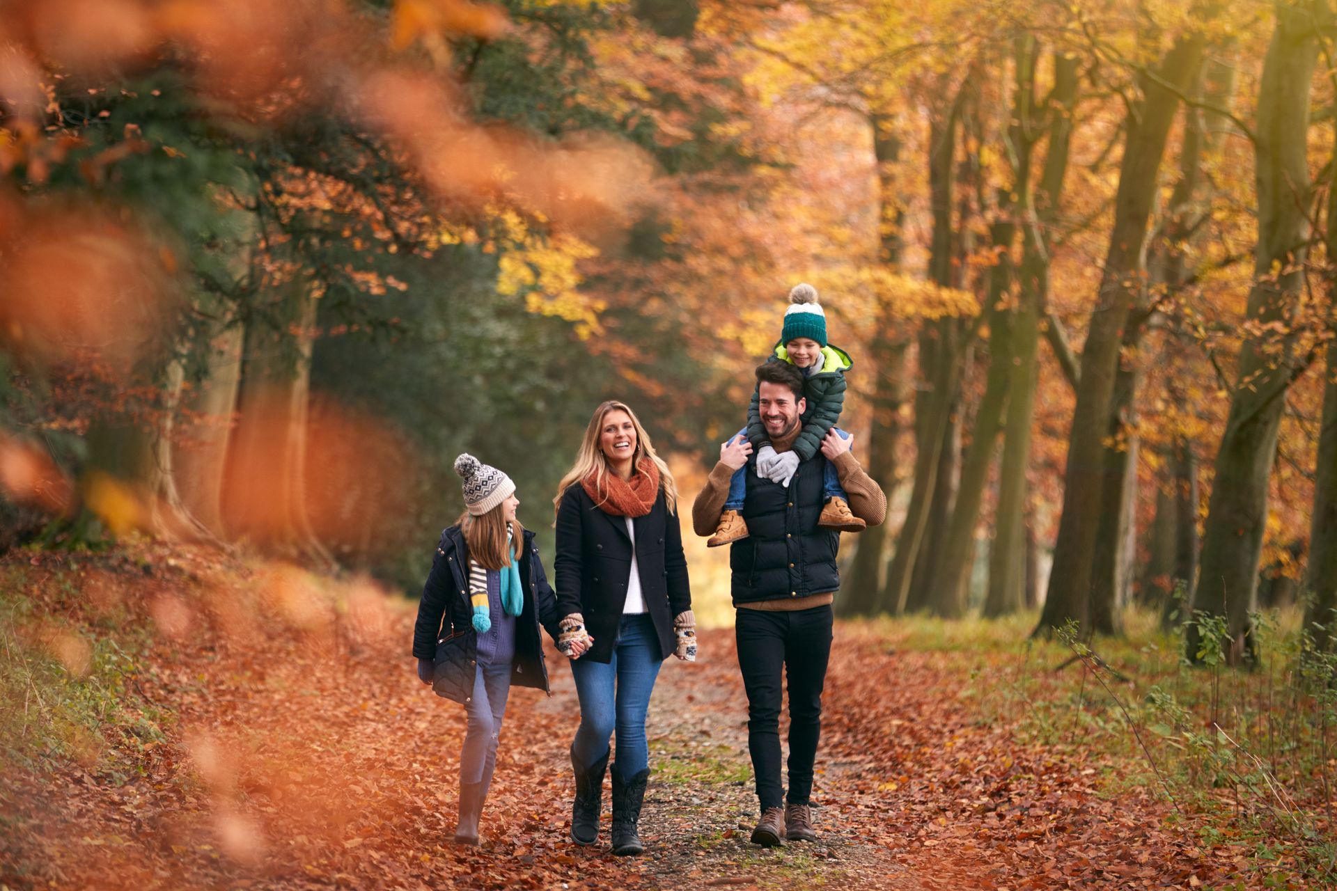A family walks through a park in autumn, with a person carrying a child on their shoulders among fall foliage.