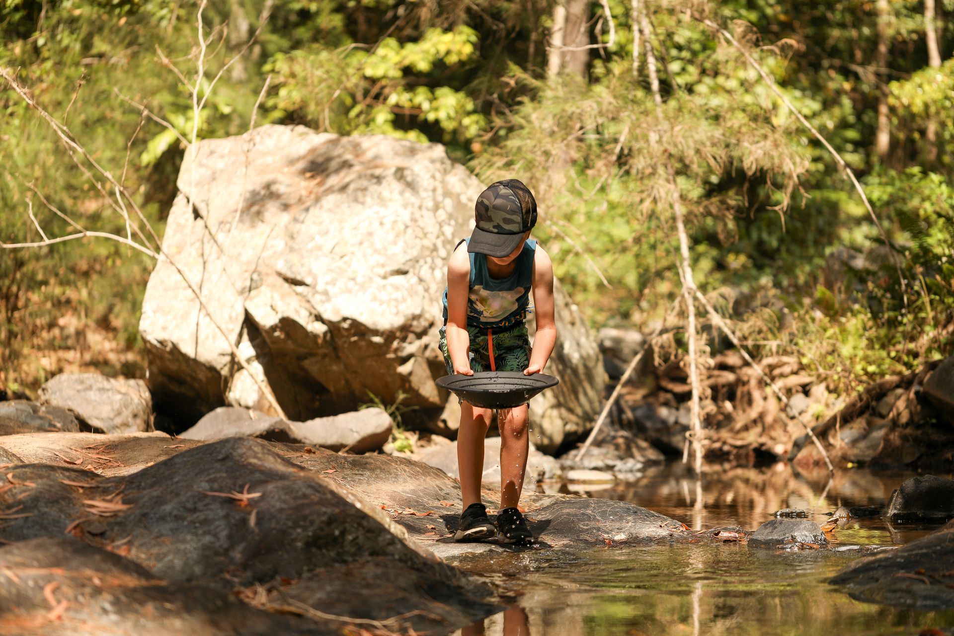 A person wearing a hat stands in a rocky stream, holding a black panning dish while looking down at the water.