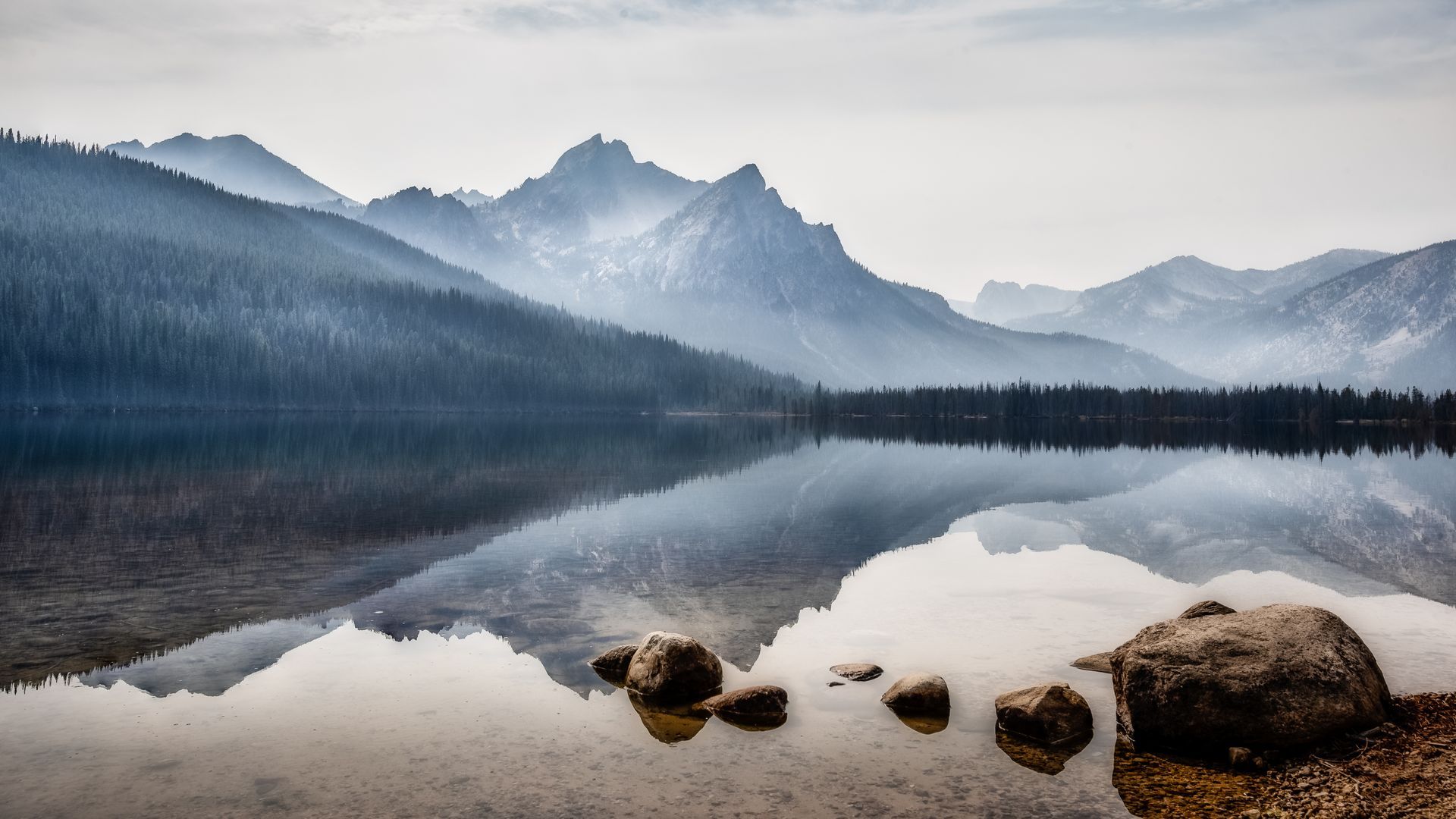 Mountains reflected in a calm lake, with a foggy sky and rocks in the foreground.