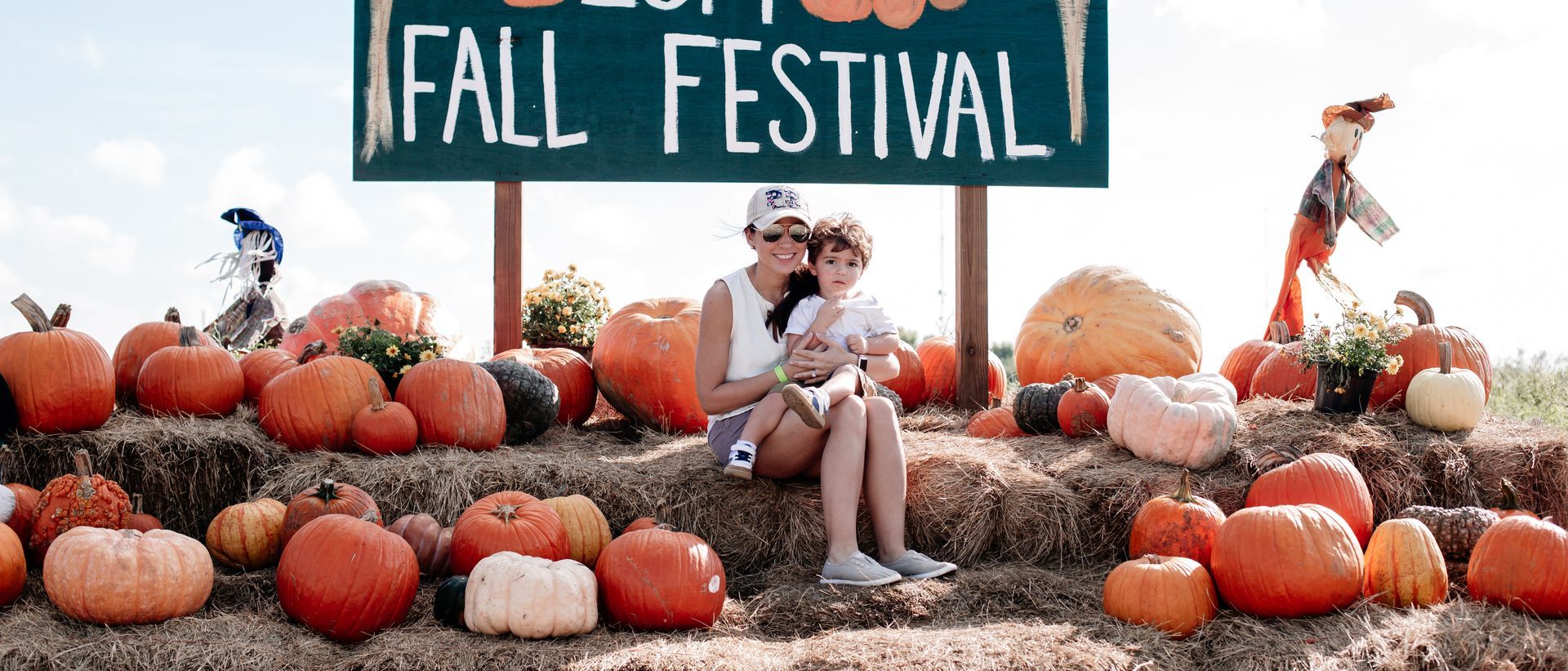 Woman and child at a fall festival. They're sitting on hay bales surrounded by pumpkins, with a festival sign.
