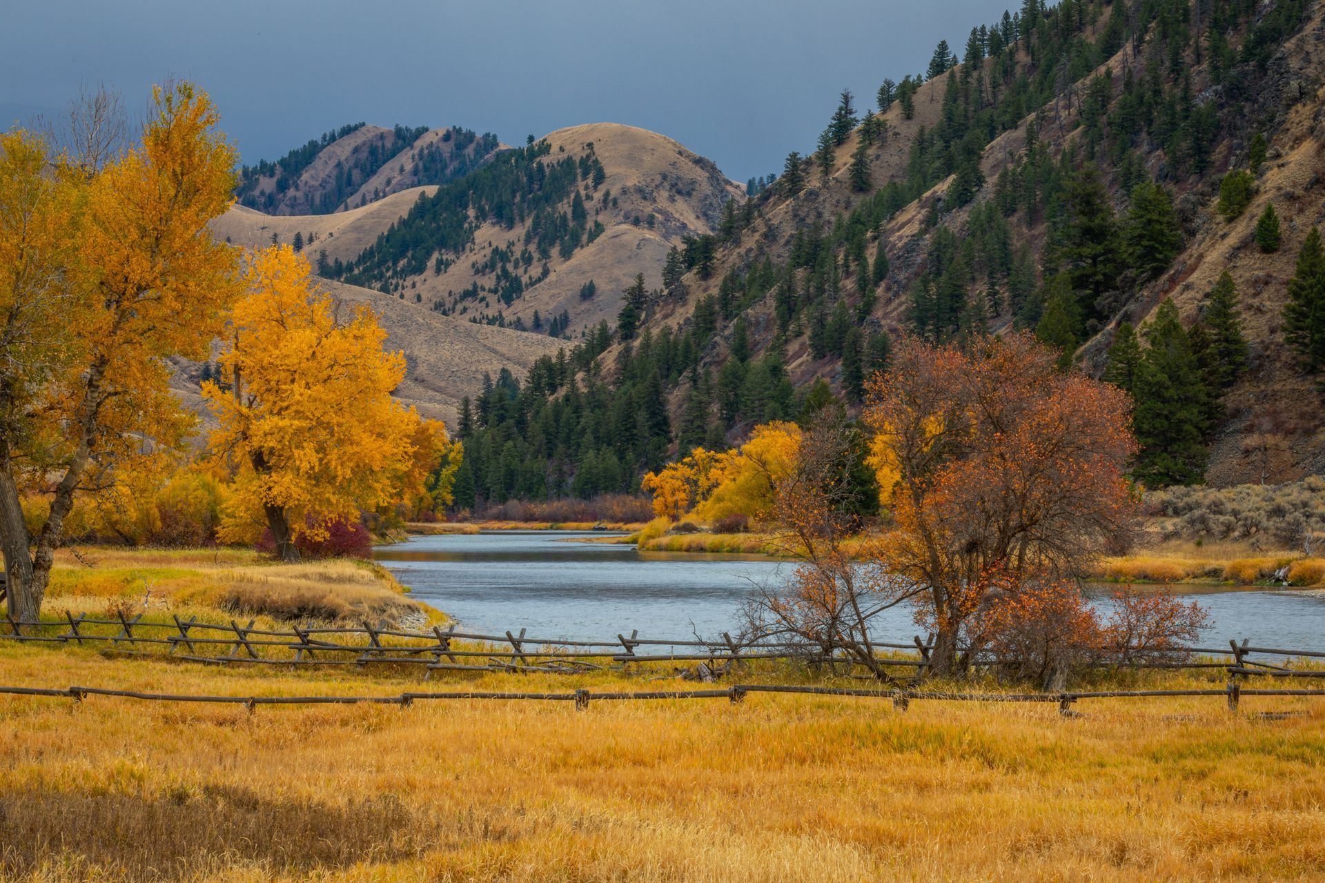 Autumnal landscape: Golden fields, river, colorful trees, wooden fence, mountains under a cloudy sky.