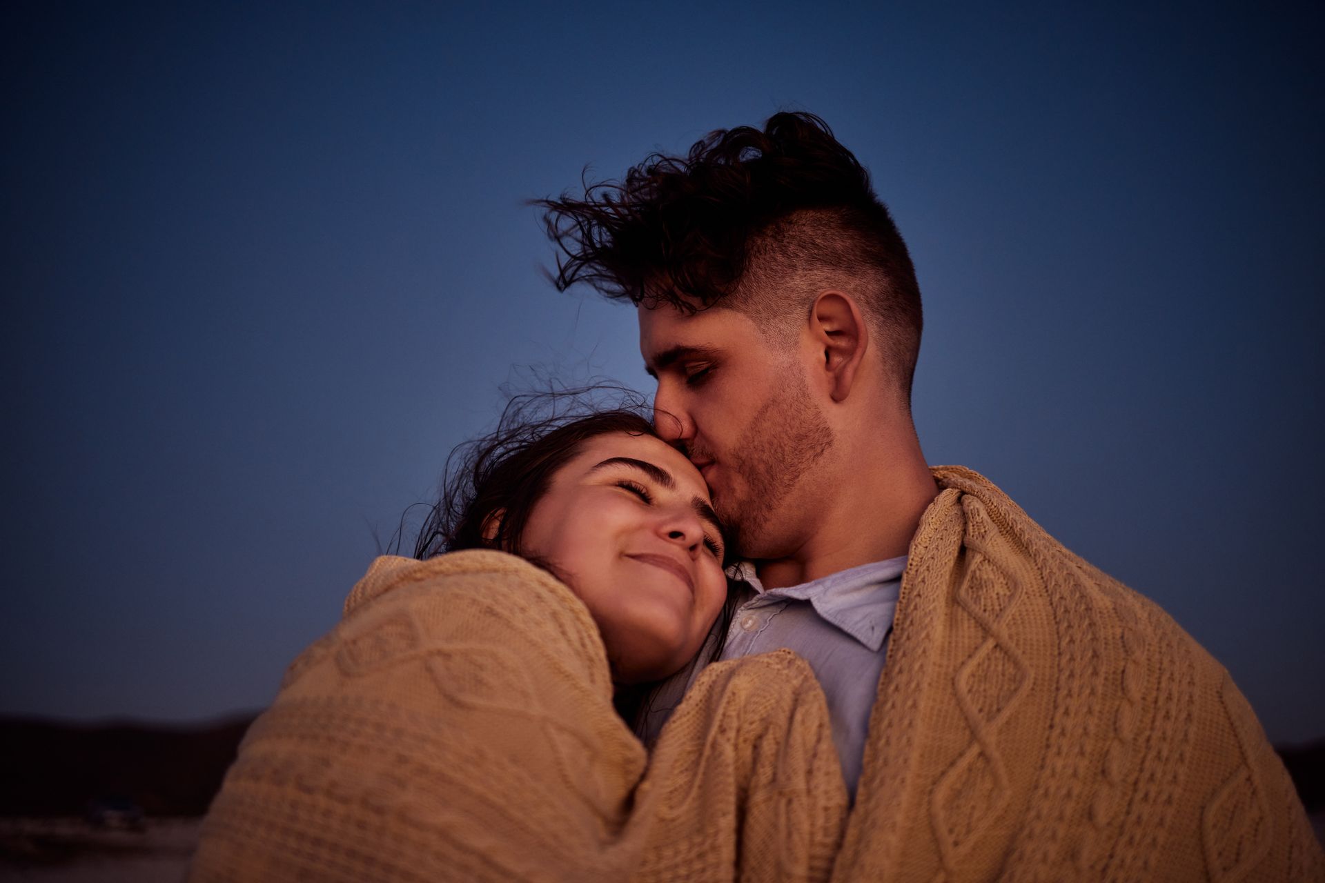 Man kisses woman's forehead as they're wrapped in a blanket, dusk sky.