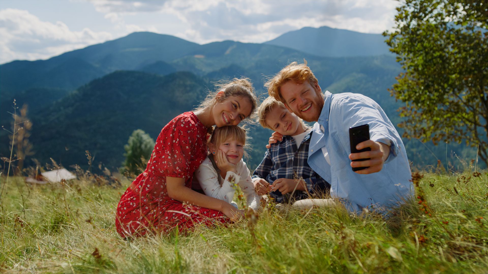 Family in a grassy field with mountains, taking a selfie. Woman in red dress, man holding phone, two children smiling.