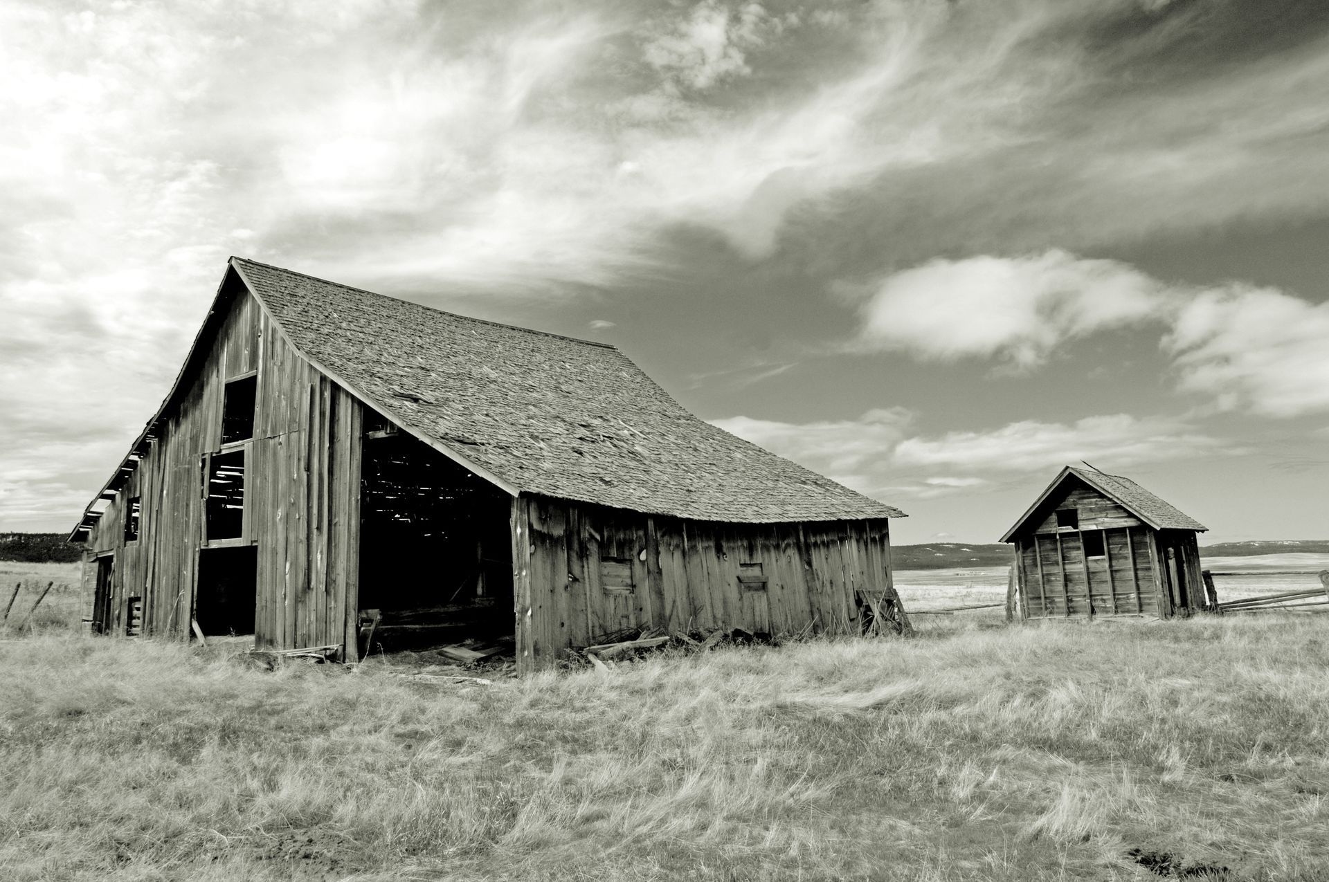 Dilapidated wooden barn and shed in a field under a cloudy sky.