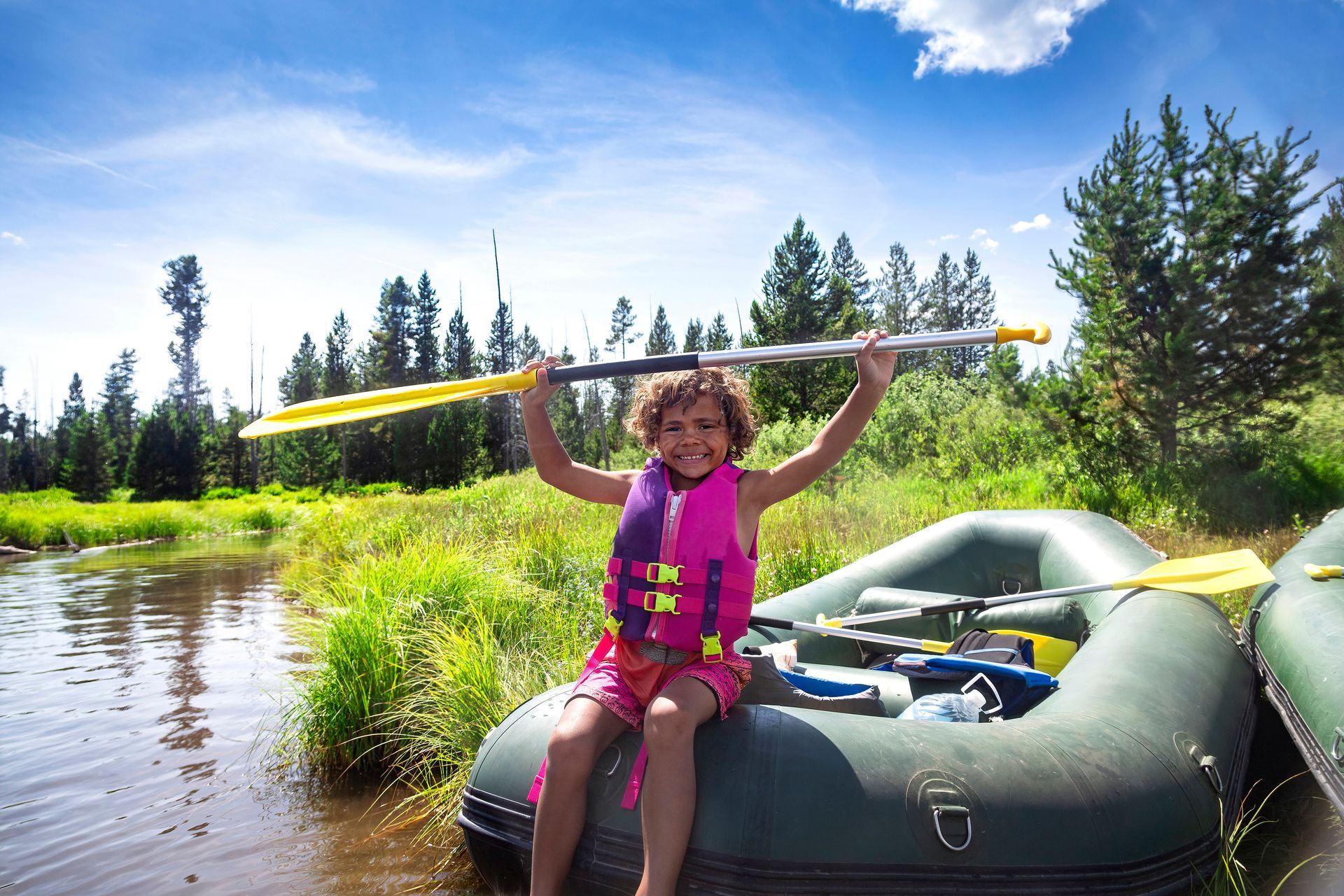 A smiling child in a pink life jacket sits on a raft in a grassy river, holding a paddle high overhead.
