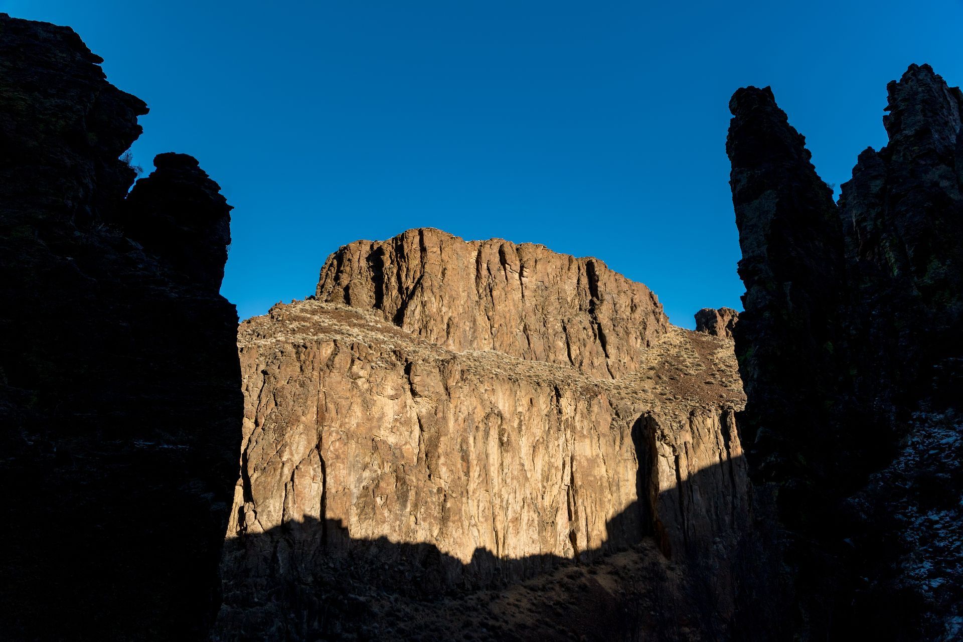 A towering rock formation, brightly lit by the sun, framed by darker rock spires, under a blue sky.